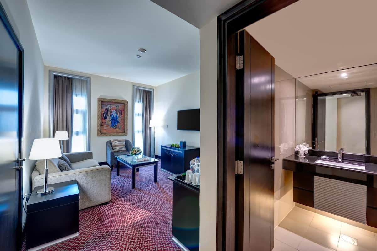 Ein Hotelzimmer mit Wohnbereich, einem Flachbildfernseher, einer Couch, einem Couchtisch, einem Esstisch und einem modernen Badezimmer mit großem Spiegel und Waschbecken.