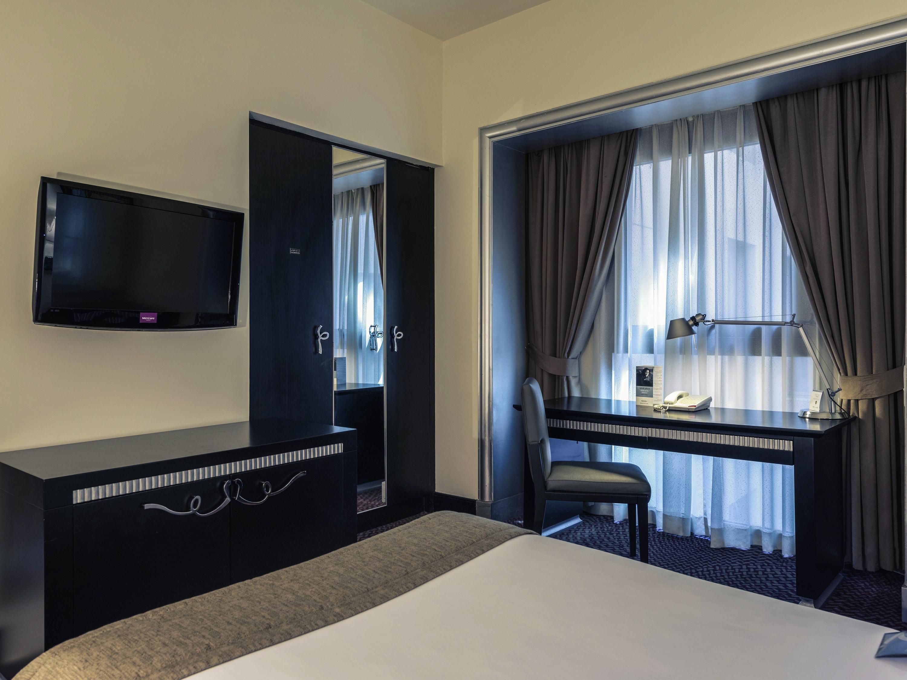 A hotel room with a bed, a desk with a chair, a television, and a window with curtains.