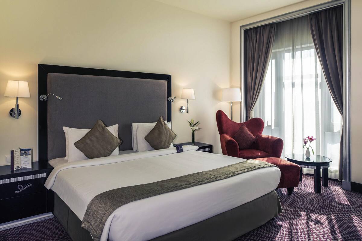 Ein Hotelzimmer mit einem großen Bett, zwei Nachttischen, einem roten Sessel, einem kleinen Tisch mit einer Vase und einem großen Fenster mit Vorhängen.