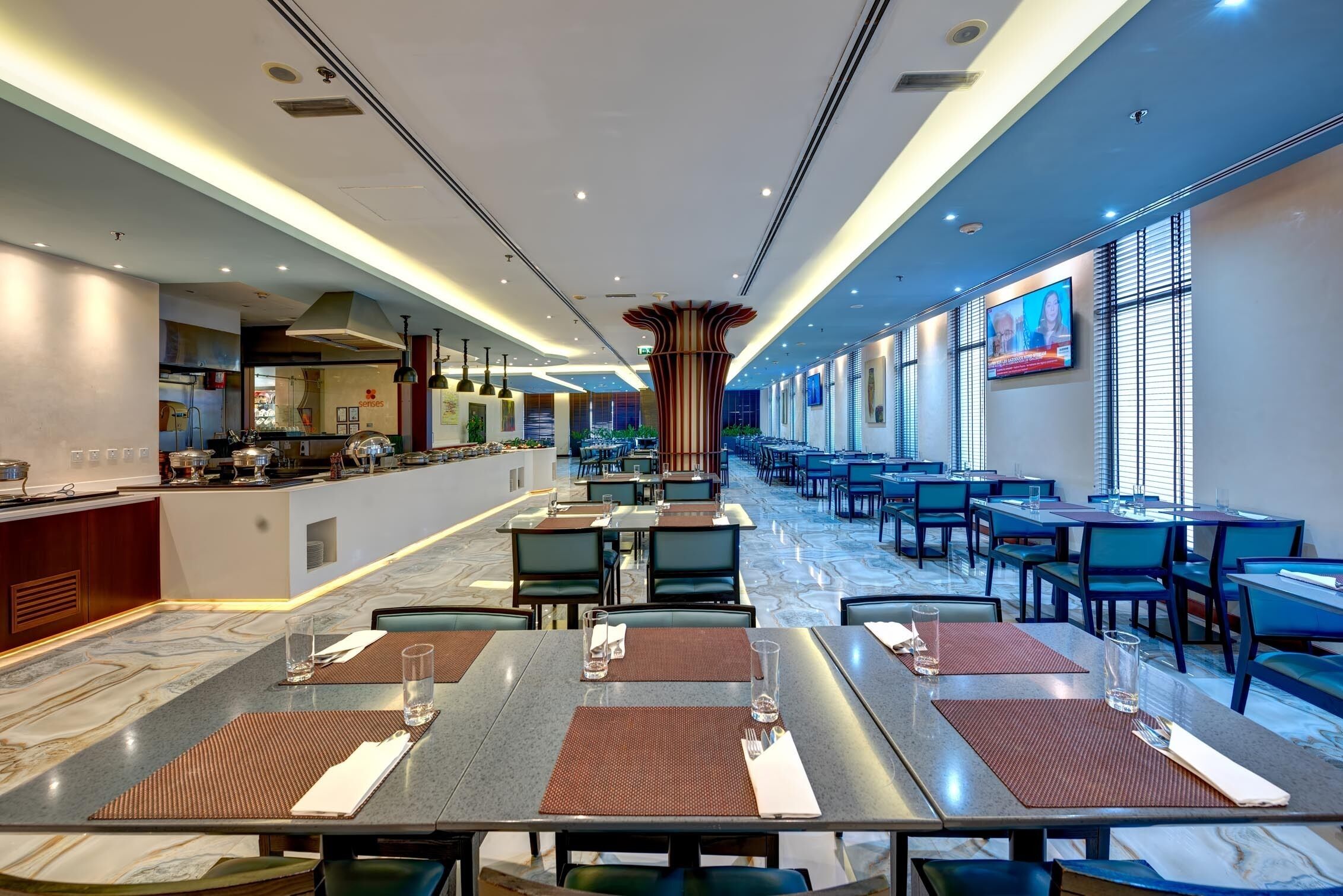 A modern hotel dining area with a long counter, multiple tables, and a television screen on the wall.