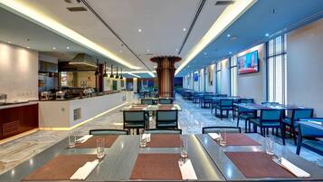 A modern hotel dining area with a long counter, multiple tables, and a television screen on the wall.