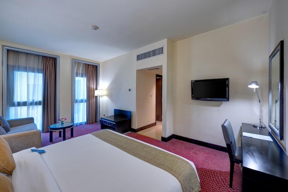 A hotel room with a large bed, a desk, a chair, a TV, and a small table with a vase.