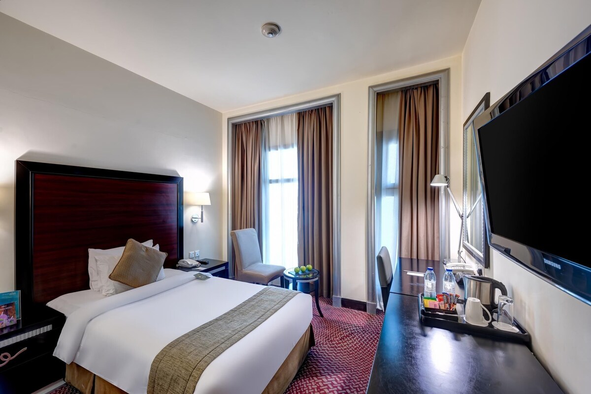 A hotel room with a large bed, a desk with a computer, a chair, a TV, and a window with curtains.