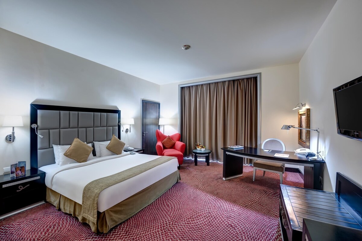 A hotel room with a large bed, a desk, a chair, a TV, and a window with curtains.