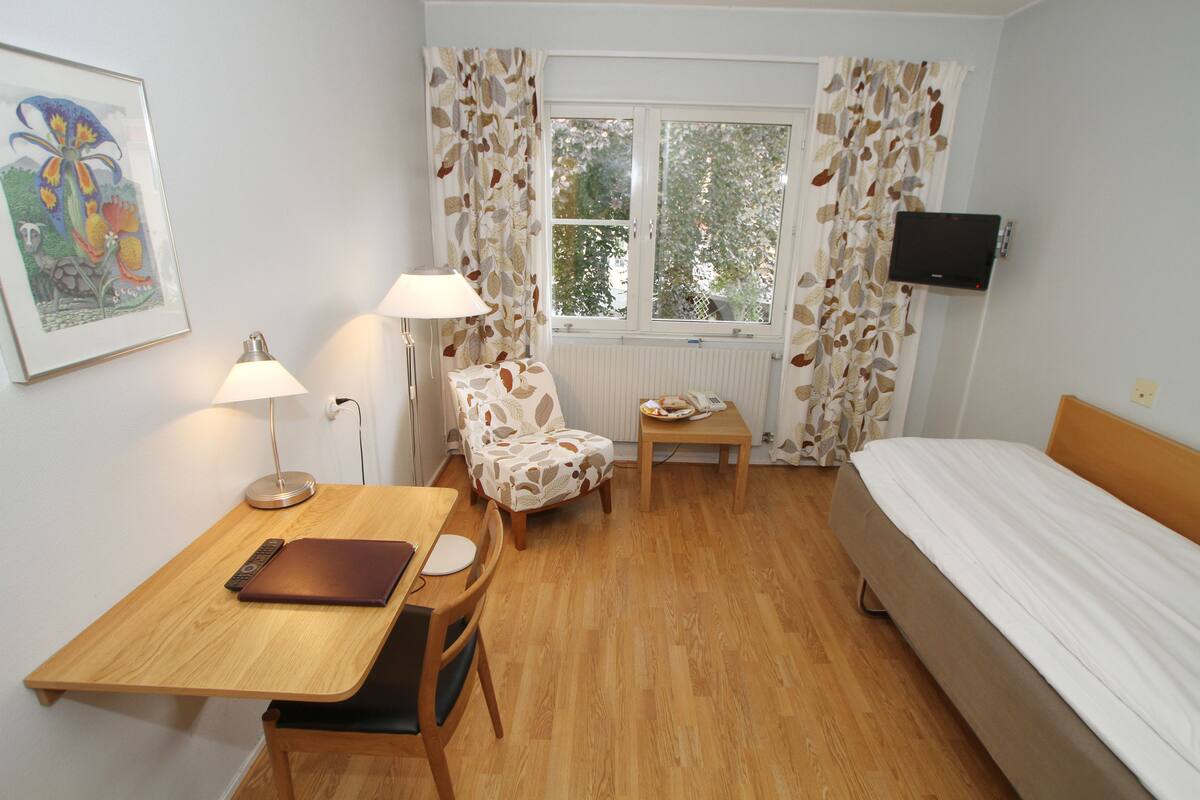 A room with a bed, a desk with a laptop, a chair, a table with a plate, a lamp, and a TV on the wall.