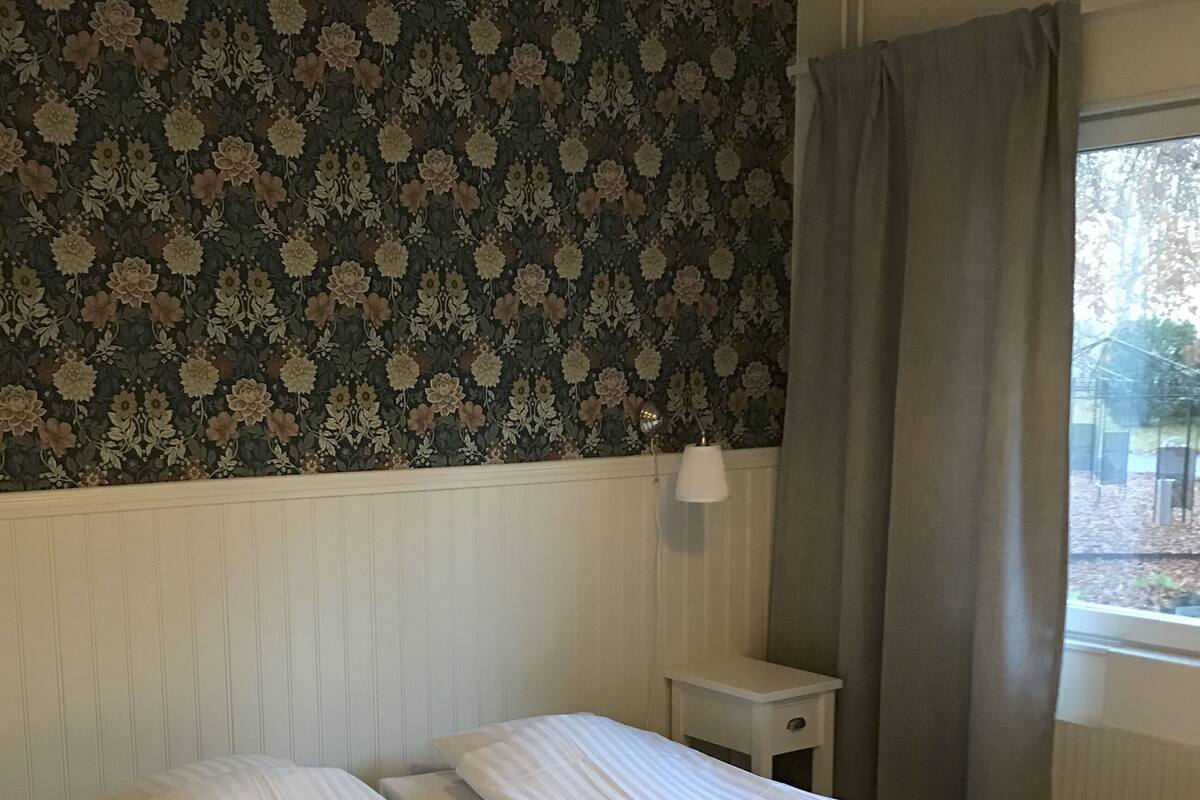 A room with two single beds, a small bedside table, a lamp, and a window with curtains.