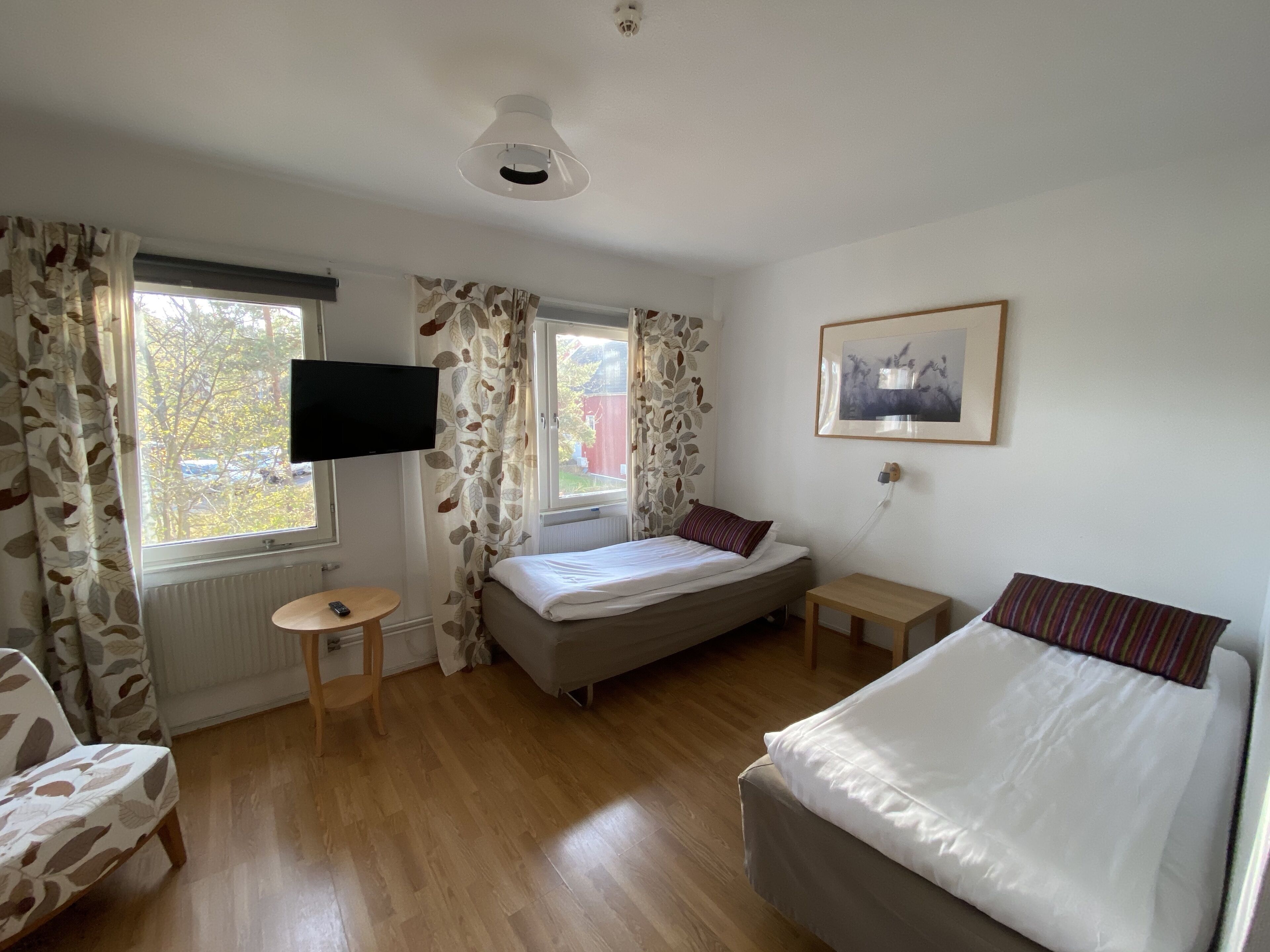 Un dormitorio con cama, televisor, una mesita, una silla y ventana con cortinas.