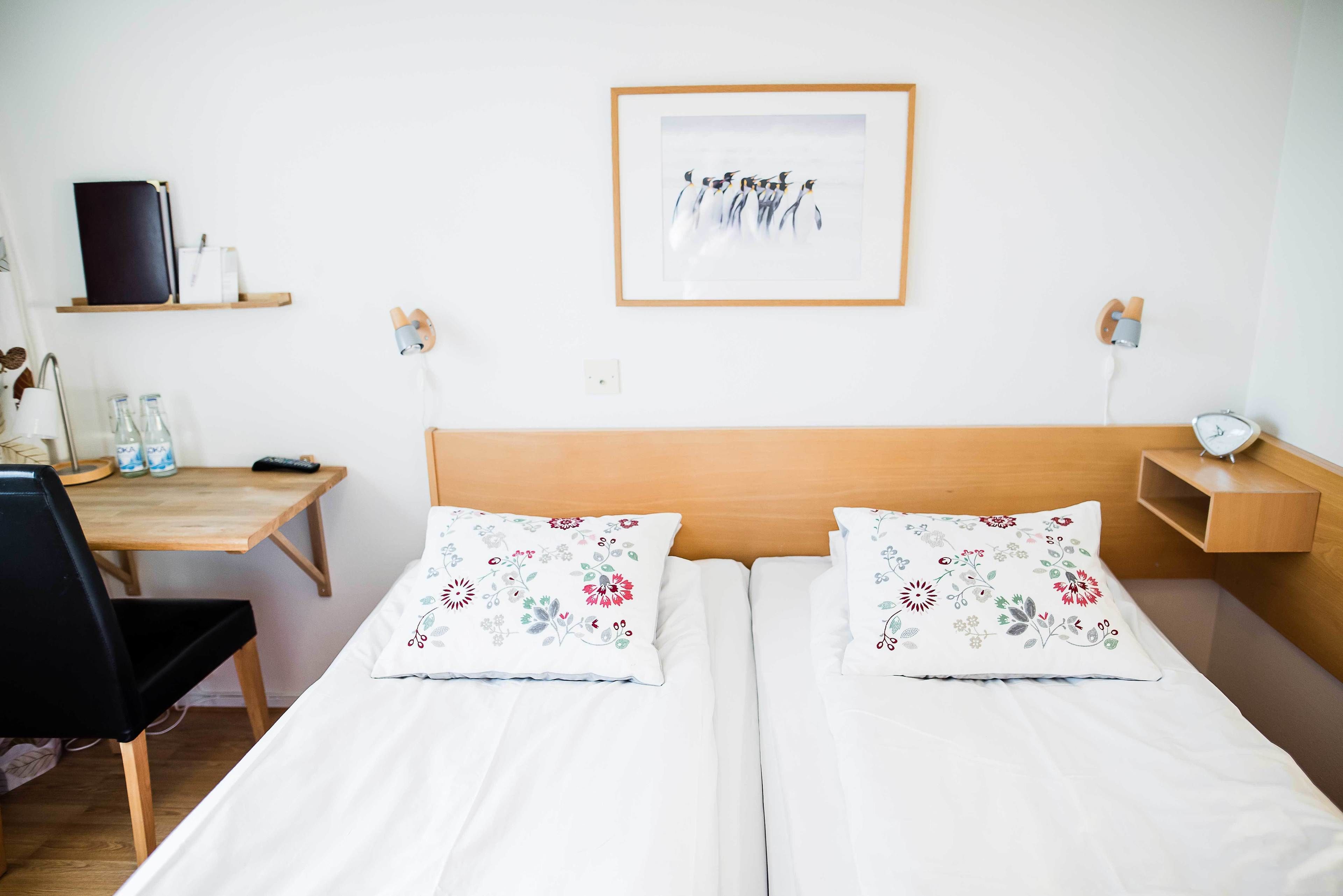 A hotel room with a wooden headboard, two single beds with floral pillows, a desk with a chair, a shelf with a book and a lamp, and a framed picture on the wall.