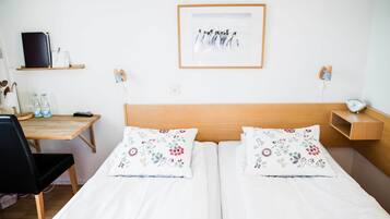 Habitación de hotel con cabecera de madera, dos camas individuales con almohadas estampadas, un escritorio con una silla, una estantería con un libro y una lámpara, y un cuadro en la pared.