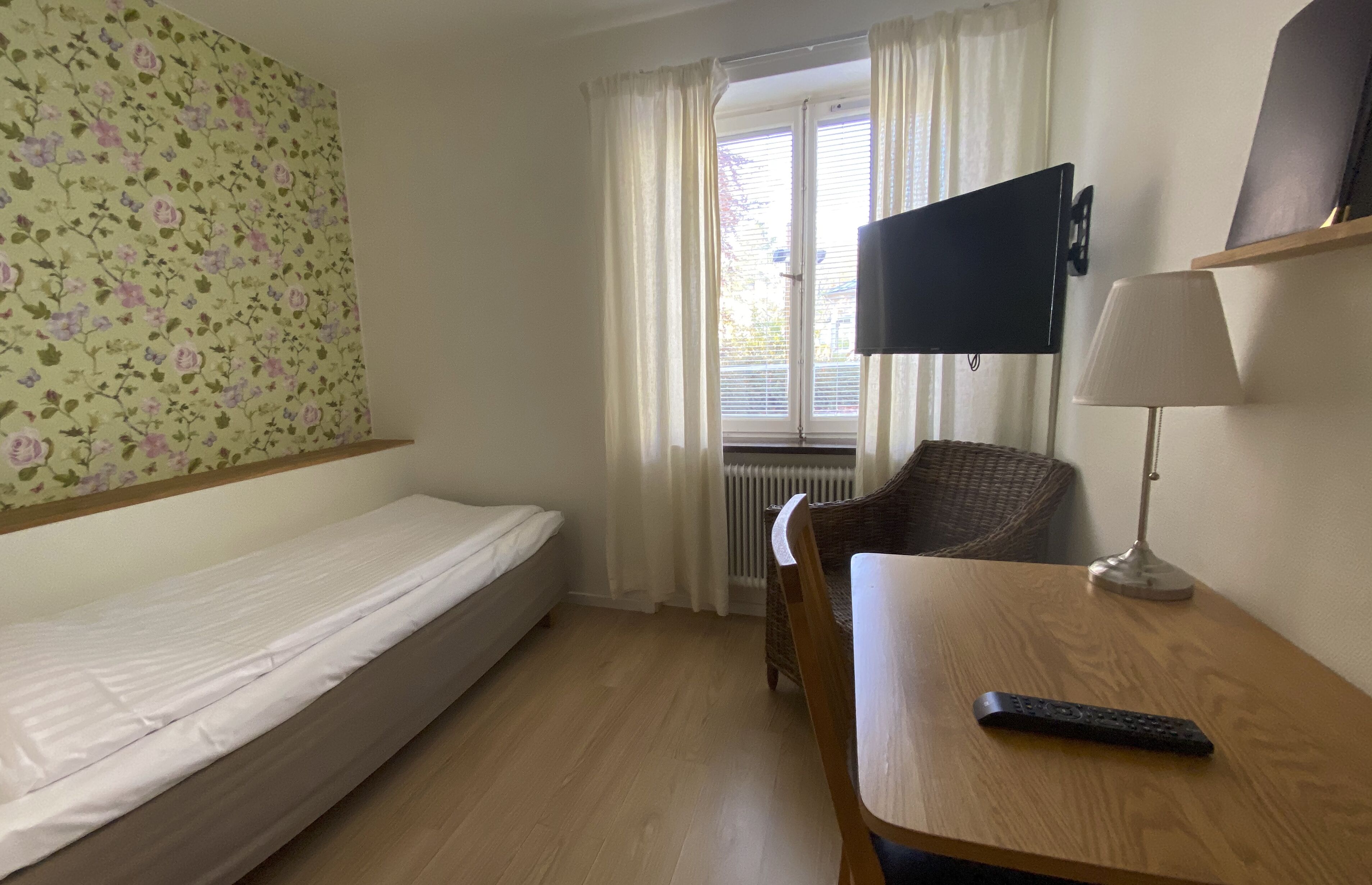 A hotel room with a single bed, a chair, a table, a TV mounted on the wall, and a window with curtains.