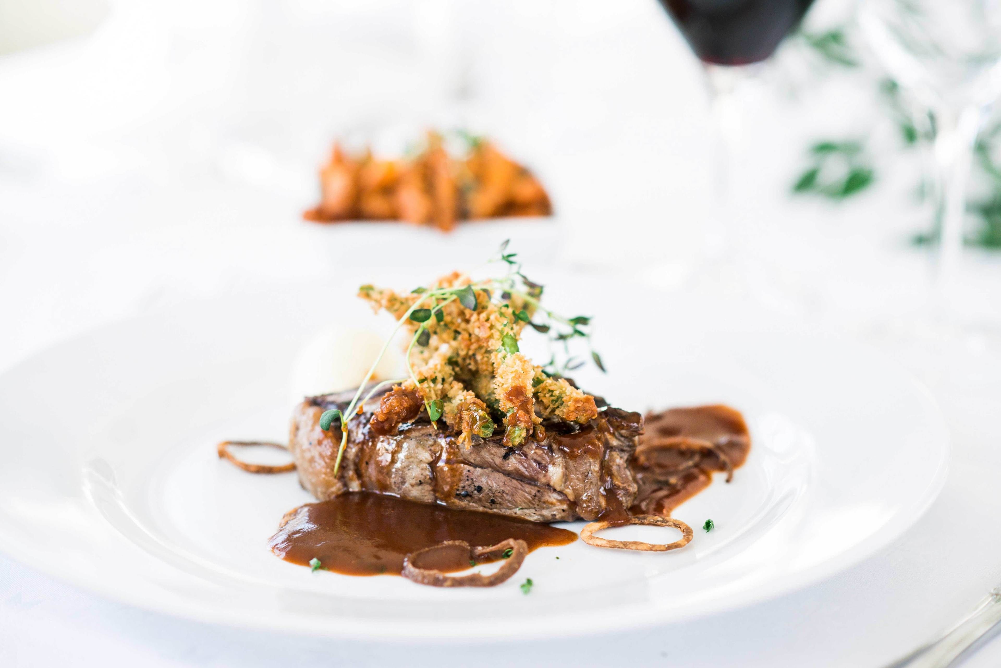 A plate of food with a steak topped with breadcrumbs and herbs, accompanied by a sauce and a small portion of mashed potatoes.