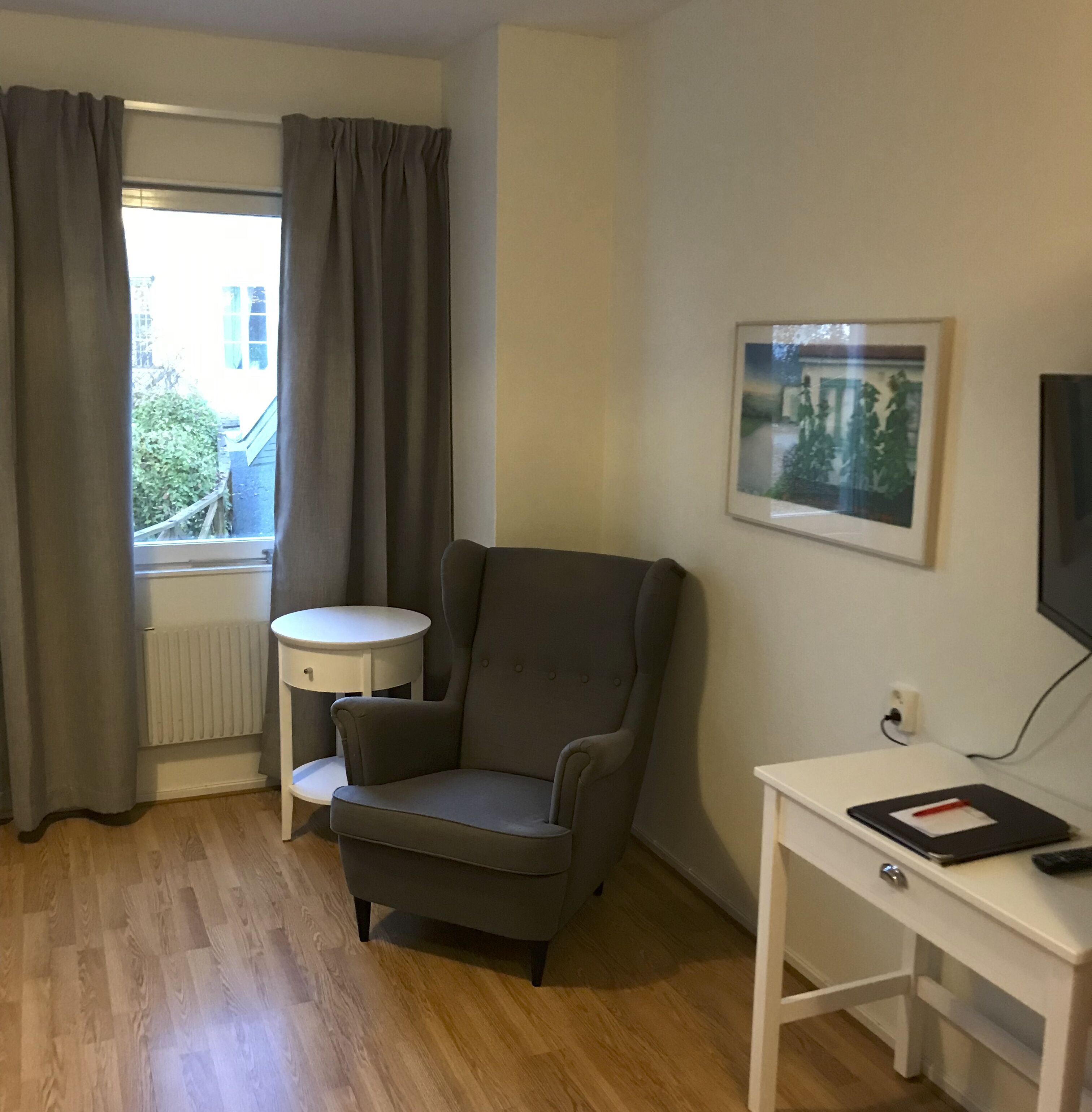 Una habitación con un sillón gris, una pequeña mesa redonda, un escritorio blanco con una computadora portátil y un control remoto, un cuadro en la pared y una ventana con cortinas grises.