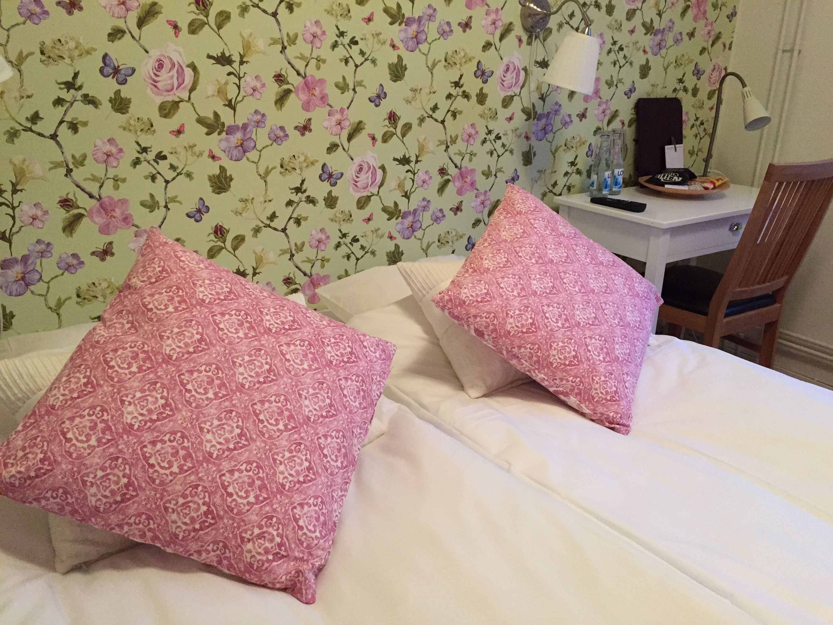Une chambre avec un papier peint à motifs floraux, deux coussins roses à motifs, une couvre-lit blanche, un bureau avec une lampe et une chaise.