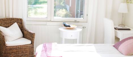 Un dormitorio con cama, una silla de mimbre, una mesita y una ventana con vistas a zonas verdes.