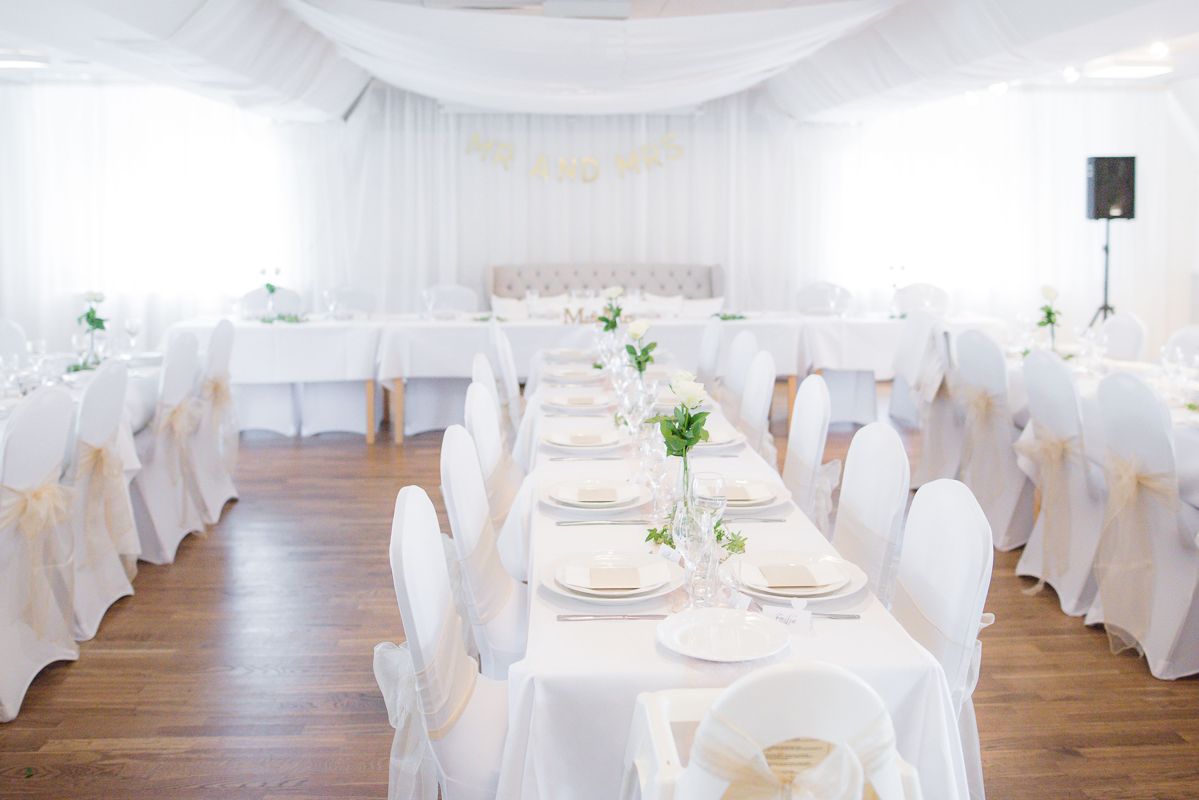 Un salón de banquetes con largas mesas dispuestas para un evento formal, con manteles blancos, sillas cubiertas de blanco y un pasillo central con arreglos florales.