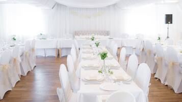 Un salón de banquetes con largas mesas dispuestas para un evento formal, con manteles blancos, sillas cubiertas de blanco y un pasillo central con arreglos florales.