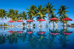 A pool area with red umbrellas, white lounge chairs, and palm trees.