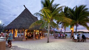 Un restaurante en la playa con cabañas de techo de paja, palmeras y una playa arenosa.