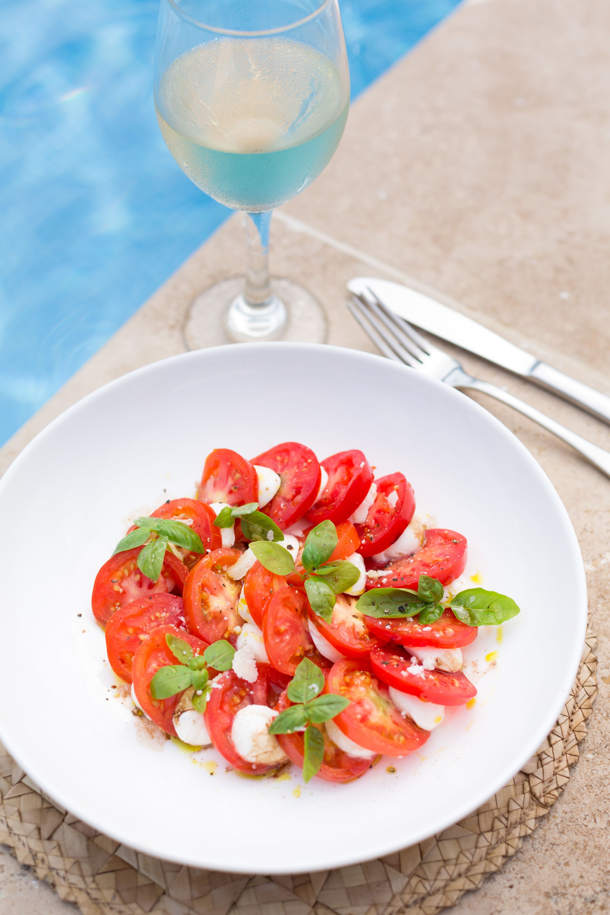 A white plate with a tomato and mozzarella salad, a glass of blue drink, and a fork and knife on a table by a pool.
