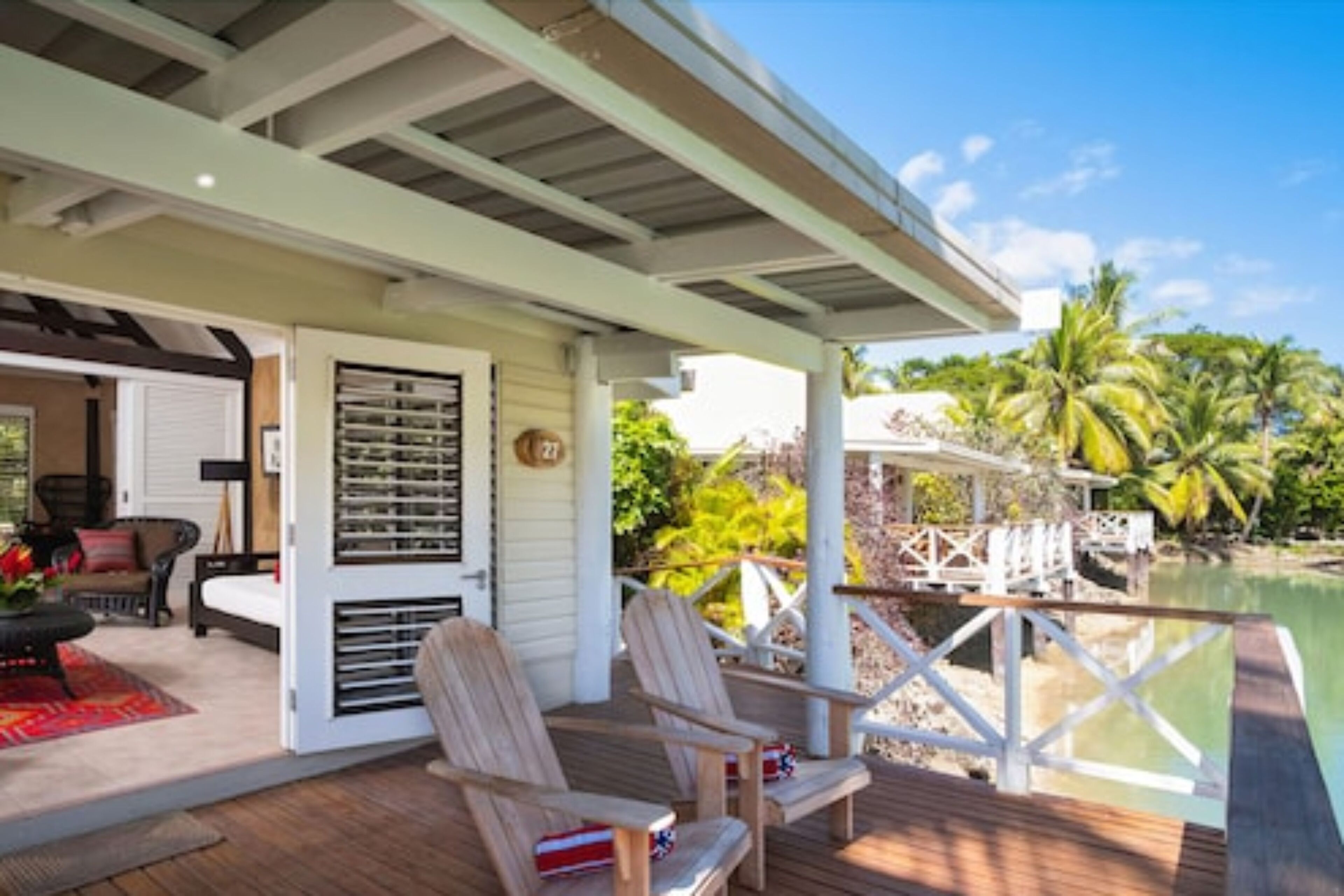 A covered deck with wooden chairs, a small table, and a view of a body of water and palm trees.
