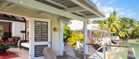 A covered deck with wooden chairs, a small table, and a view of a body of water and palm trees.