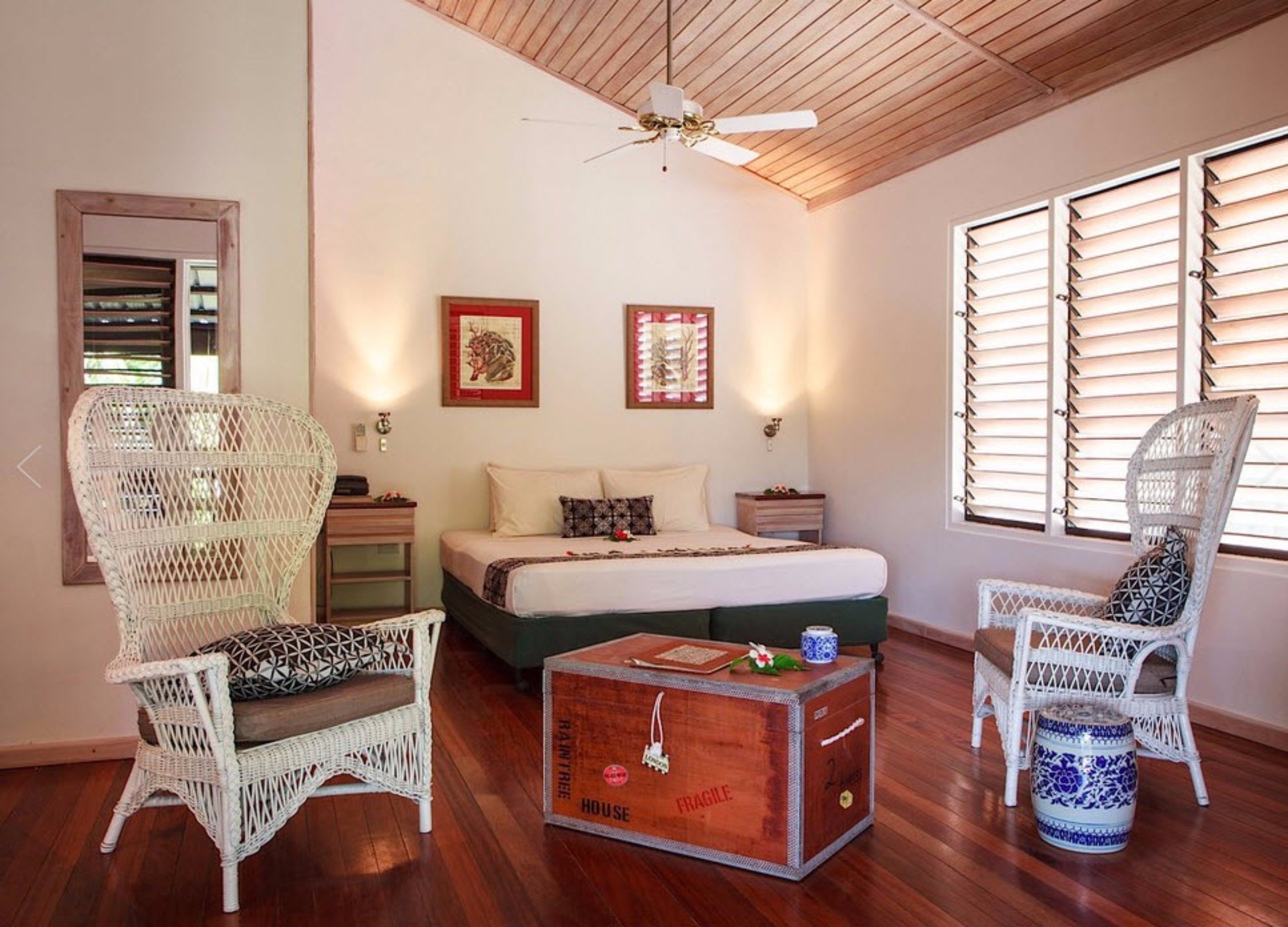 A bedroom with a bed, two nightstands, a wooden ceiling fan, wicker chairs, a wooden chest of drawers, and a window with blinds.