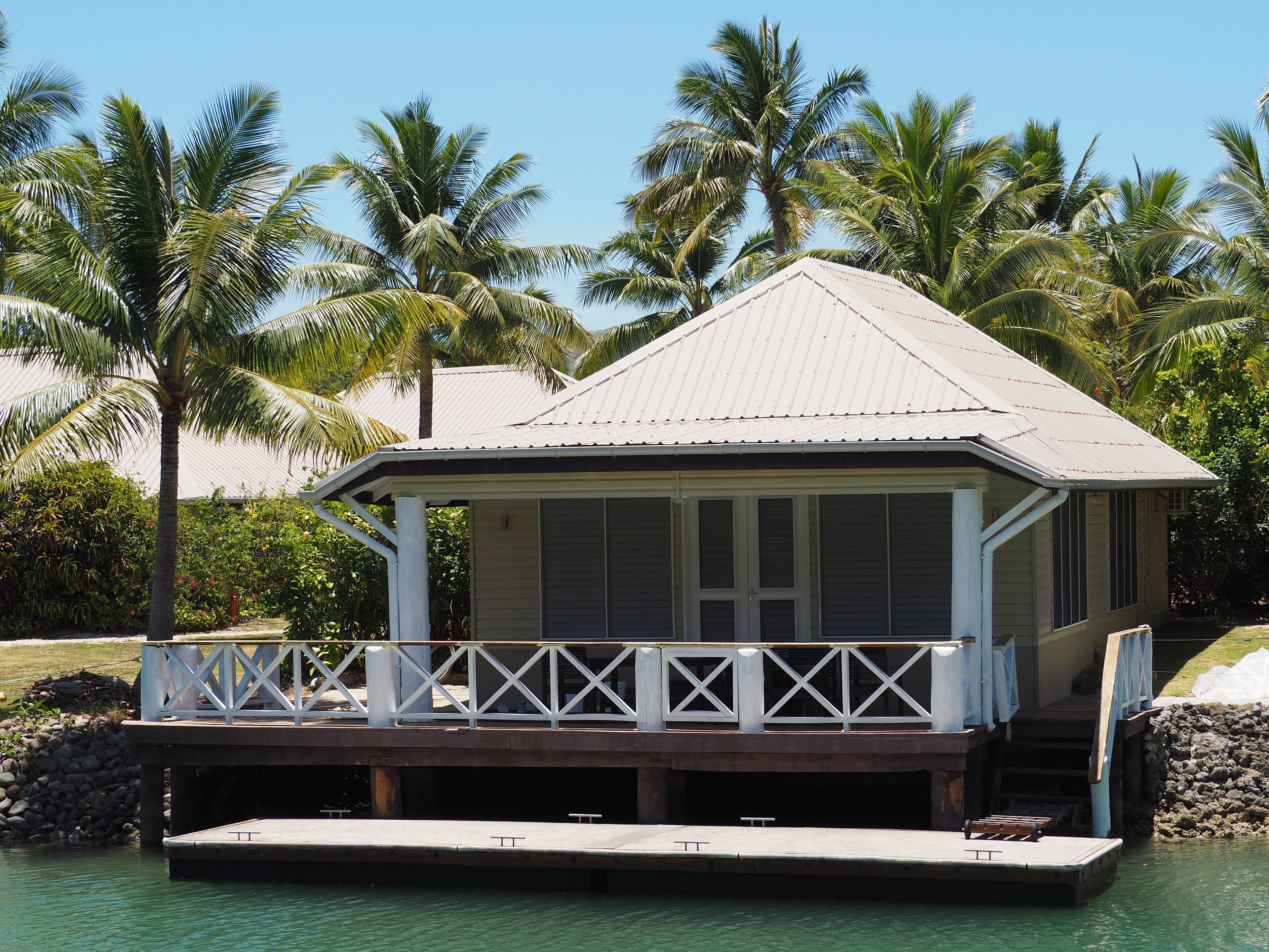 A small, single-story building with a white exterior and a metal roof, situated on a dock over water, surrounded by palm trees.