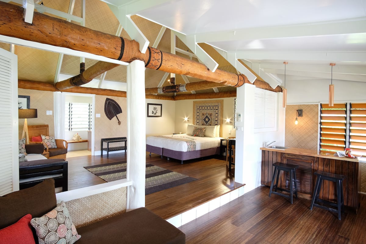 Une pièce spacieuse avec un grand lit, un plafond en bois, un coin cuisine avec des tabourets et un coin salon avec un canapé.
