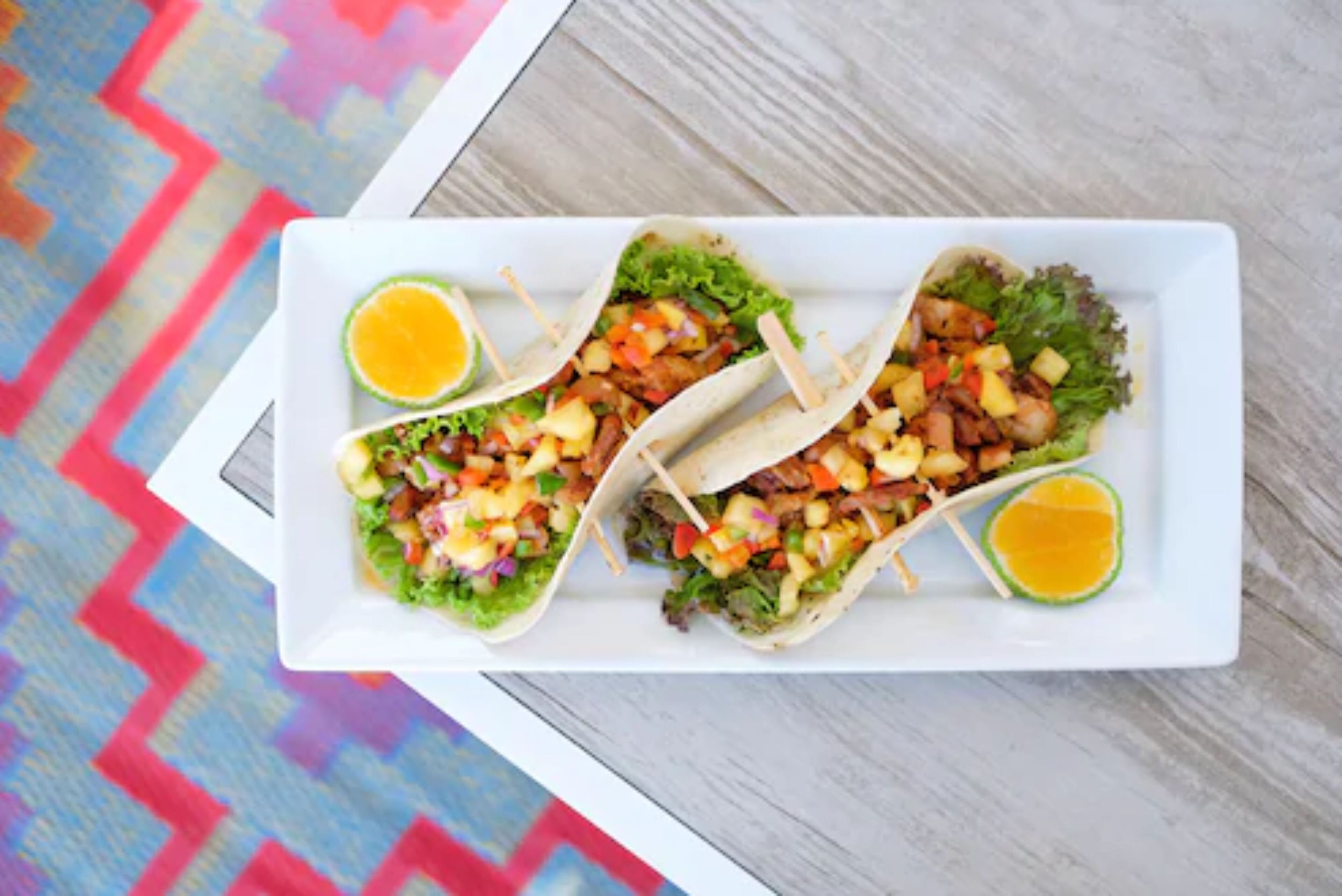 Un plato blanco rectangular con tres tacos sobre un tapete tejido colorido.