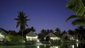 Un resort tranquilo con bungalows sobre el agua, una piscina apacible y palmeras bajo un cielo nocturno.