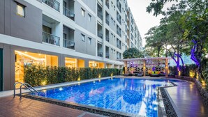 Una piscina con plataforma de cerámica, rodeada de un jardín ajardinado y un edificio de varios pisos con balcones.