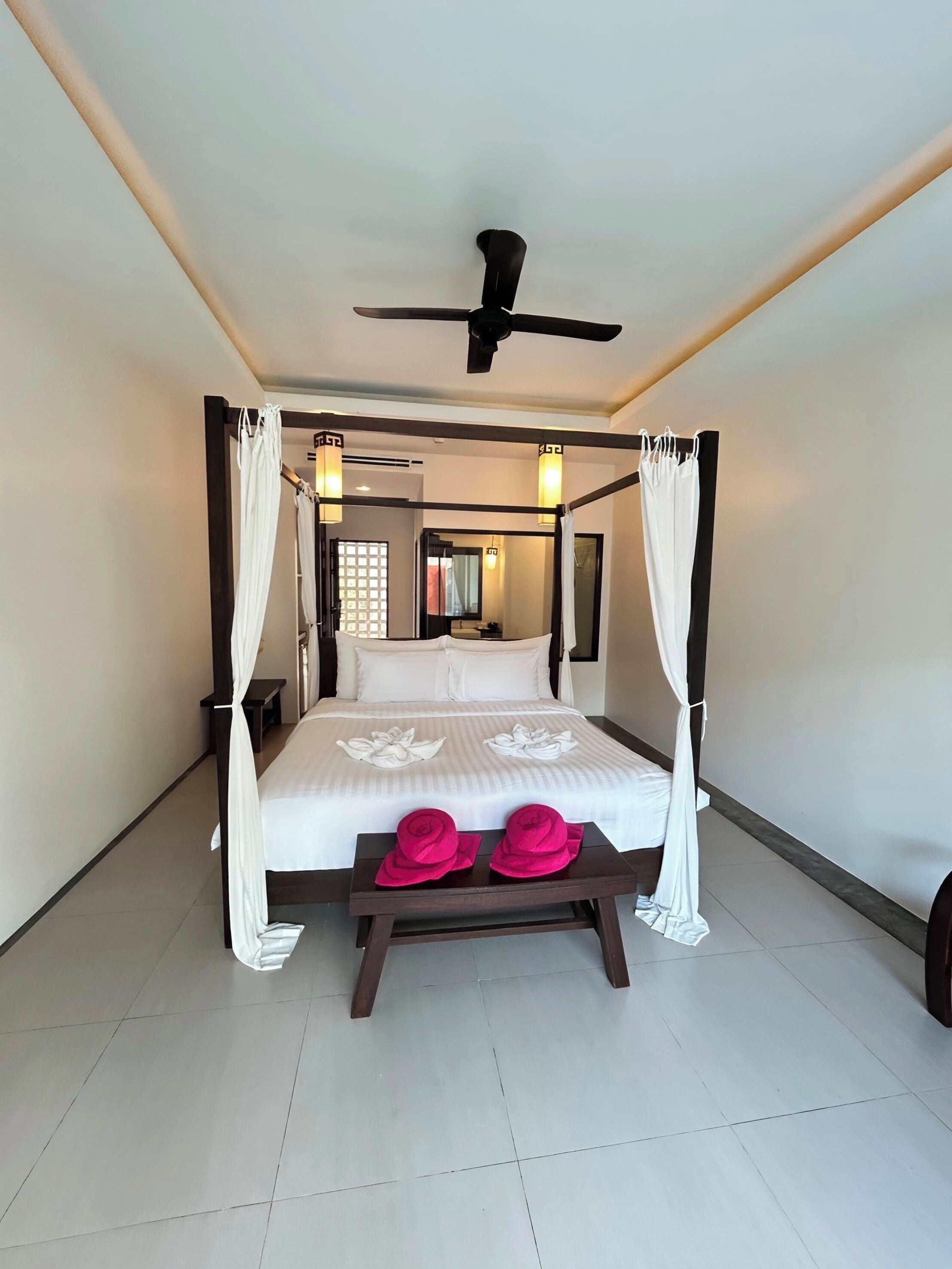 Una cama con dosel, ropa de cama blanca, un ventilador de techo y una mesita de noche con sombreros rosas.