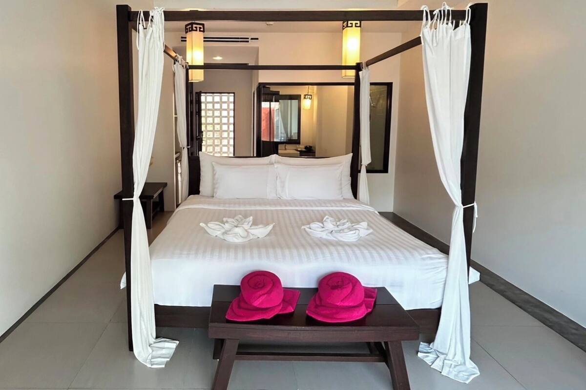 Una cama con dosel y cortinas de encaje, con ropa de cama blanca y una disposición de toallas con motivos florales. Un banco de madera con dos sombreros rosas está frente a la cama.