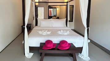 Una cama con dosel y cortinas de encaje, con ropa de cama blanca y una disposición de toallas con motivos florales. Un banco de madera con dos sombreros rosas está frente a la cama.