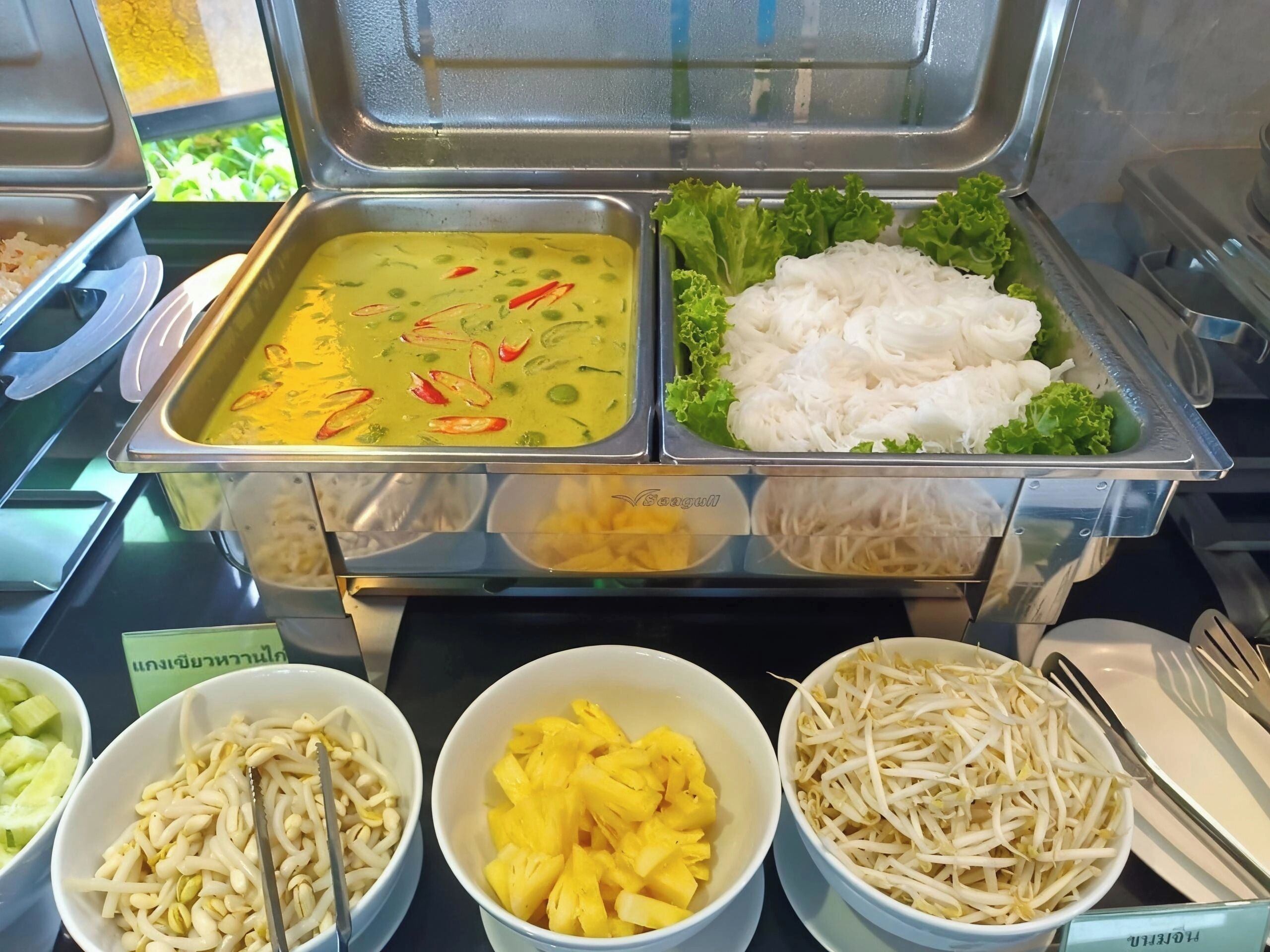 Un bufé con diversos platos, incluyendo un curry verde, fideos blancos y verduras frescas.