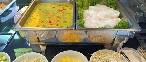 Un bufé con diversos platos, incluyendo un curry verde, fideos blancos y verduras frescas.