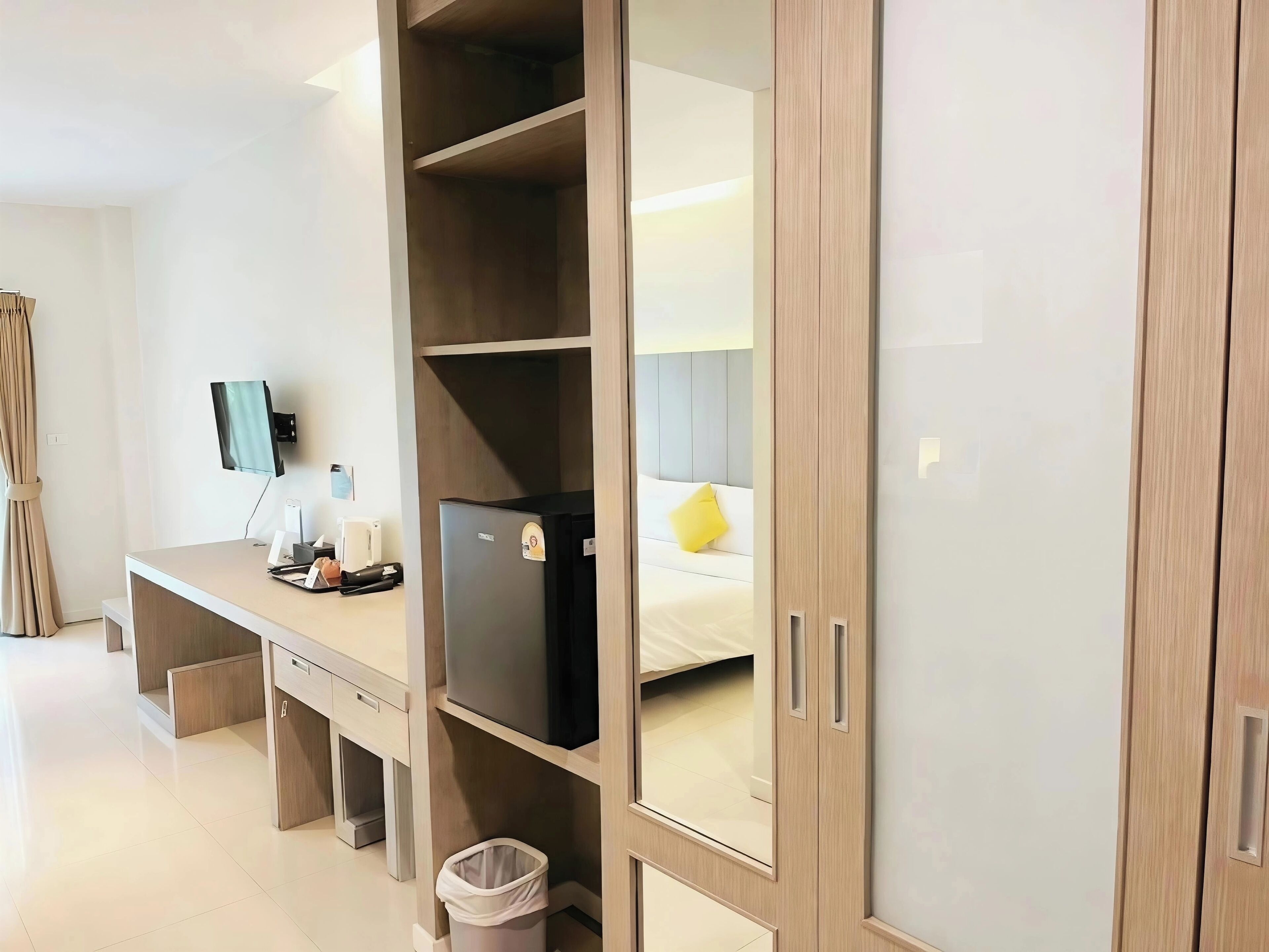 A hotel room with a wooden desk, a small refrigerator, a trash bin, and a bed with a yellow pillow.
