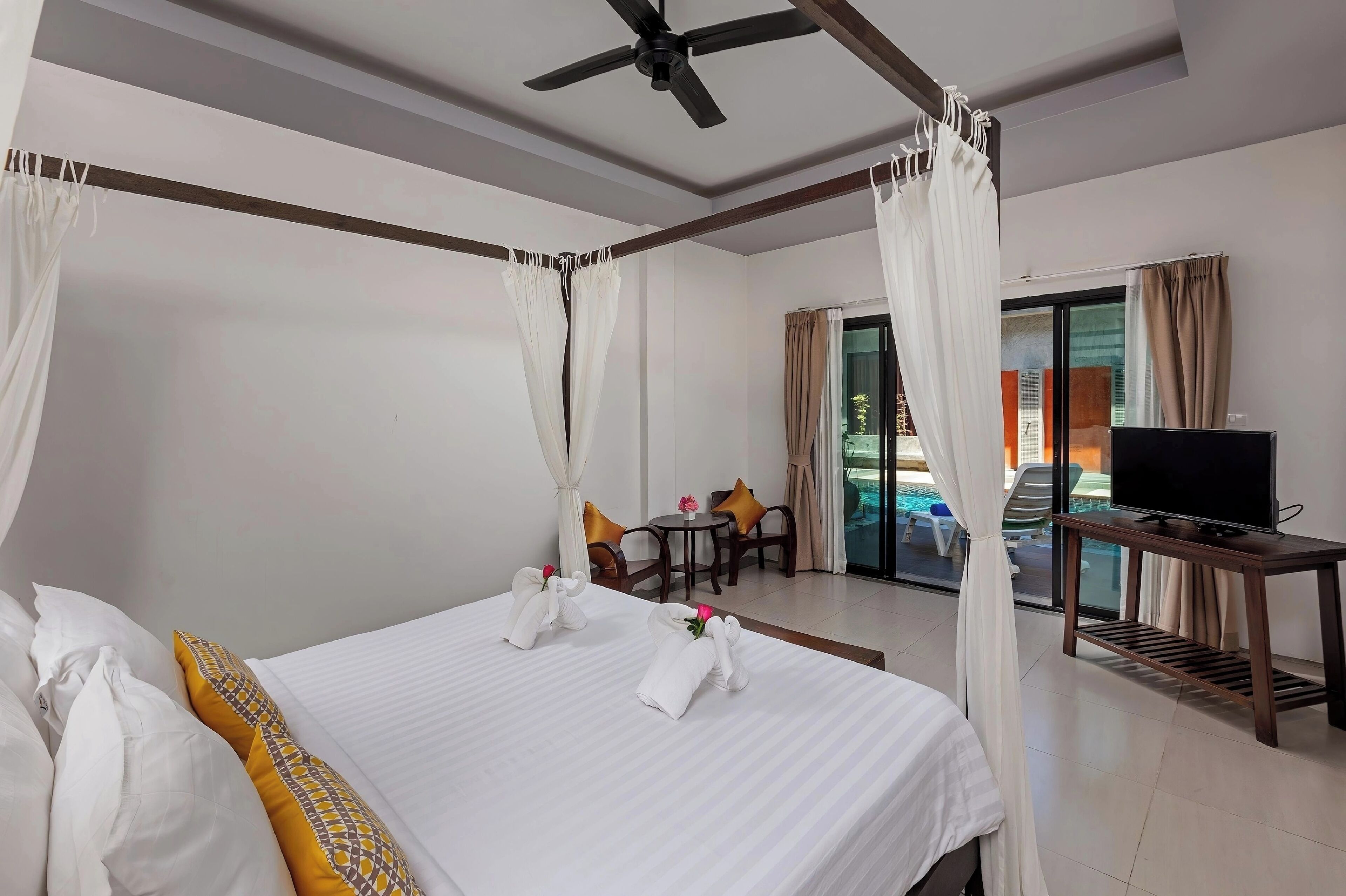Habitación de hotel con cama, ventilador de techo, televisor sobre un soporte y vista al área de la piscina.