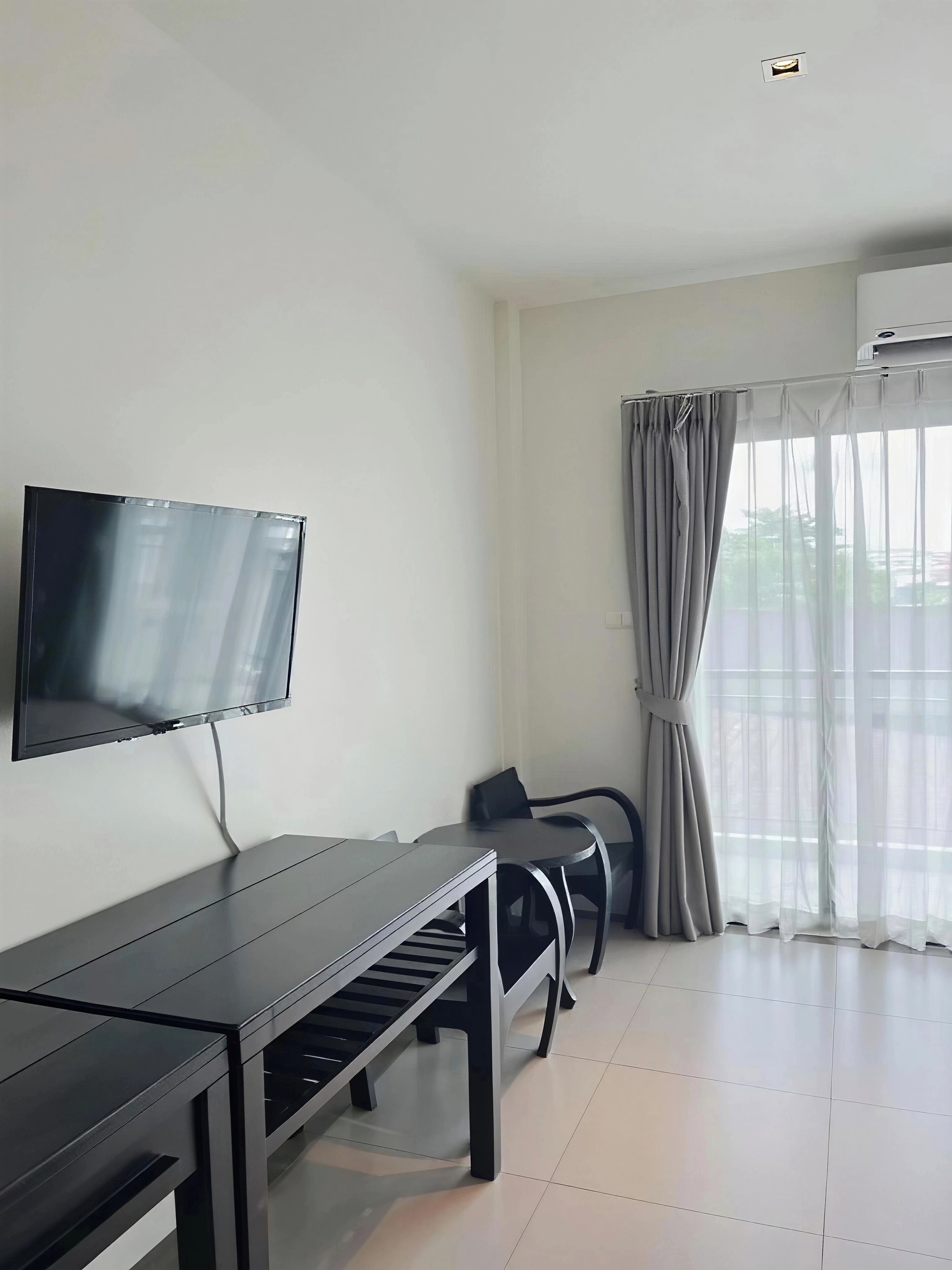 A modern room with a flat-screen TV mounted on the wall, a dark wooden table, two black chairs, and a large window with sheer curtains.