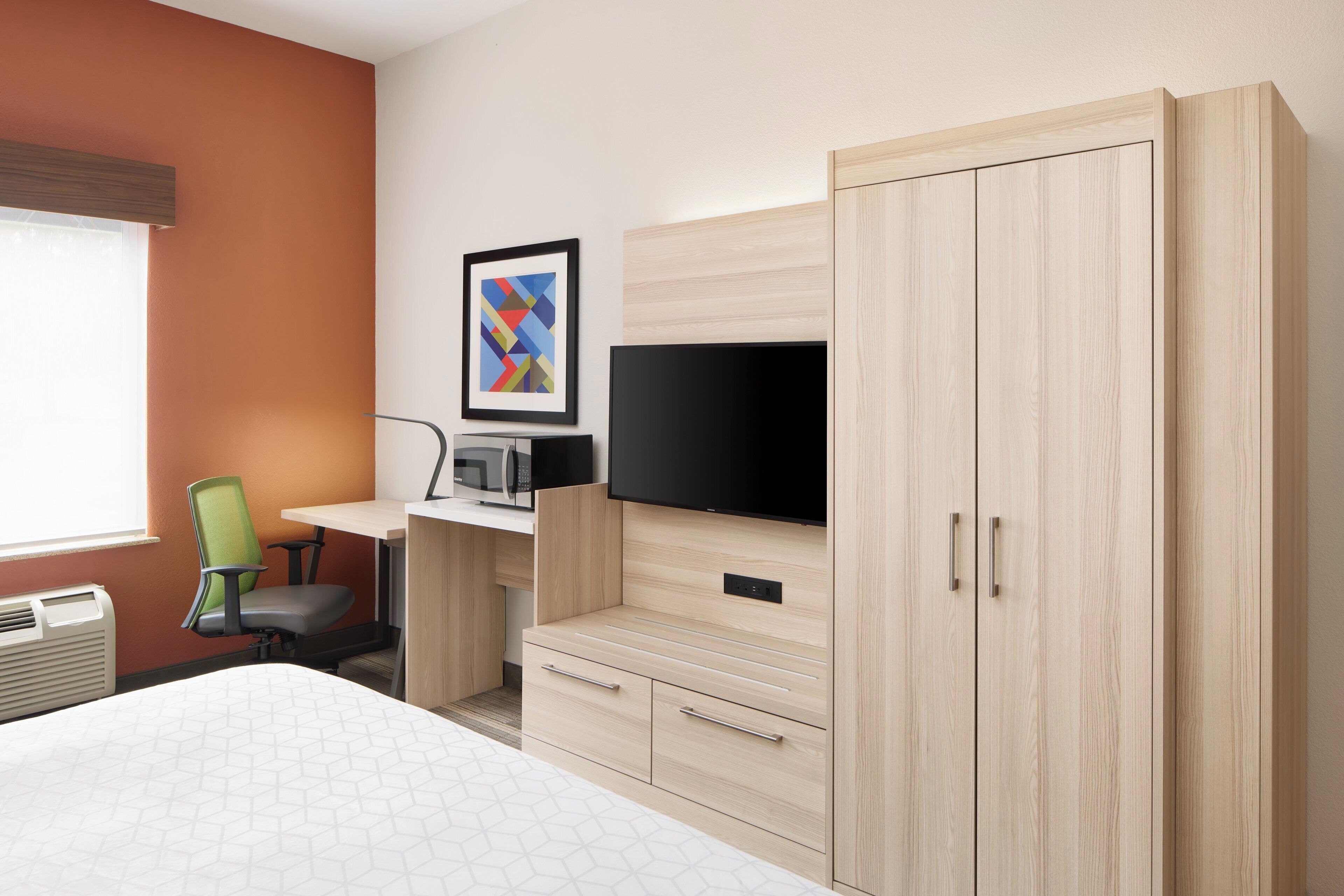A hotel room with a bed, a desk with a computer and a flat-screen TV, a chair, and a wardrobe.