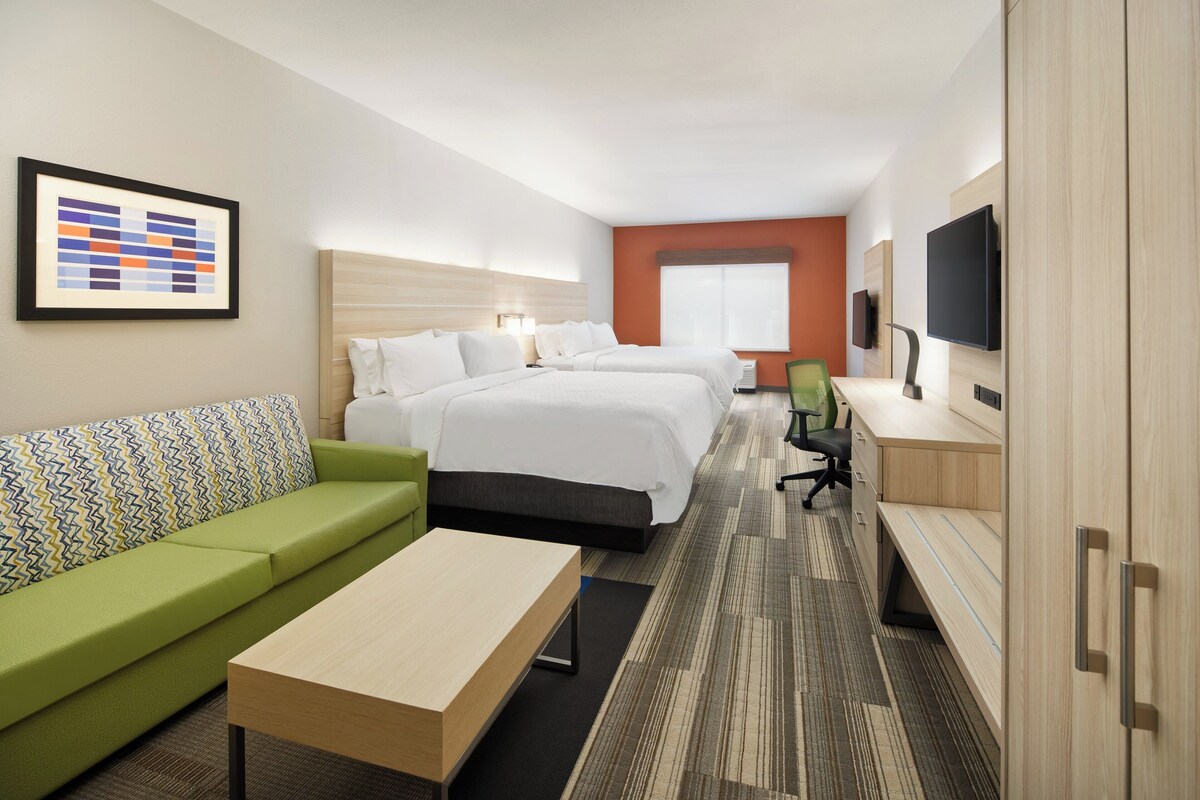 Ein Hotelzimmer mit zwei Betten, einem grünen Sofa, einem Holztisch, einem Fernseher und einem gerahmten Bild an der Wand.