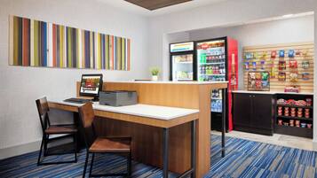 A hotel room with a desk, chairs, a vending machine, and a snack bar.