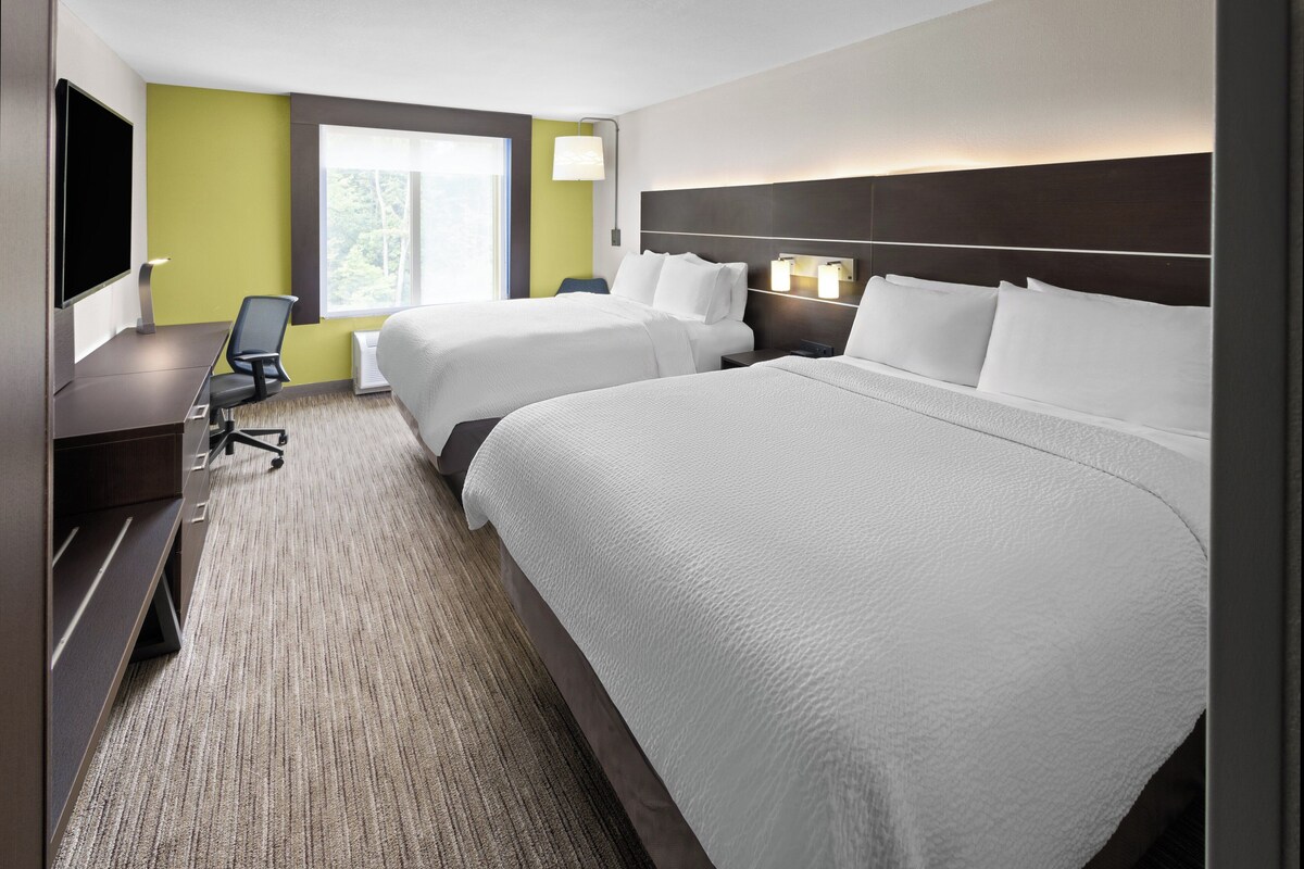 A hotel room with two beds, a desk, a chair, a TV, and a window with a view of trees.