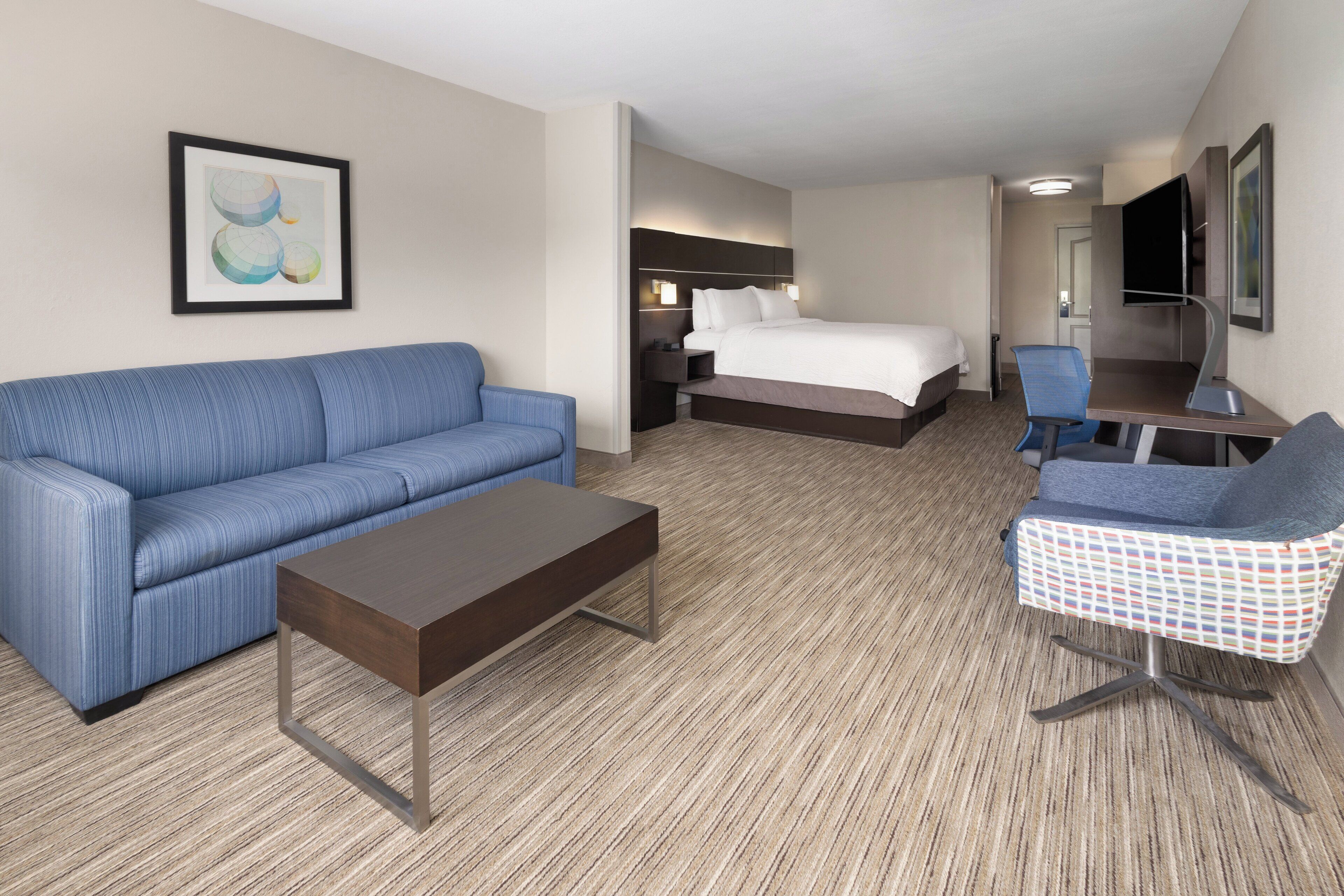 A hotel room with a blue sofa, a small table, a bed with a white comforter, a desk with a chair, and a TV mounted on the wall.