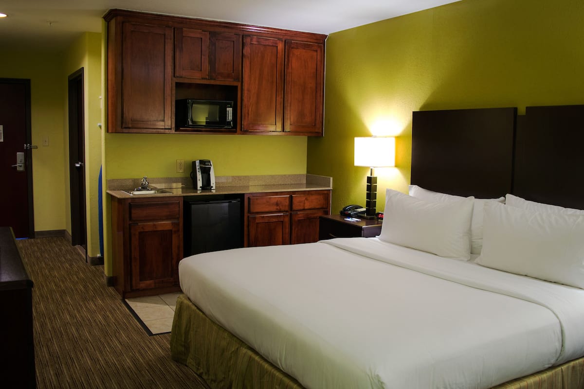 A hotel room with a bed, a desk, a microwave, a coffee maker, and a nightstand with a lamp.