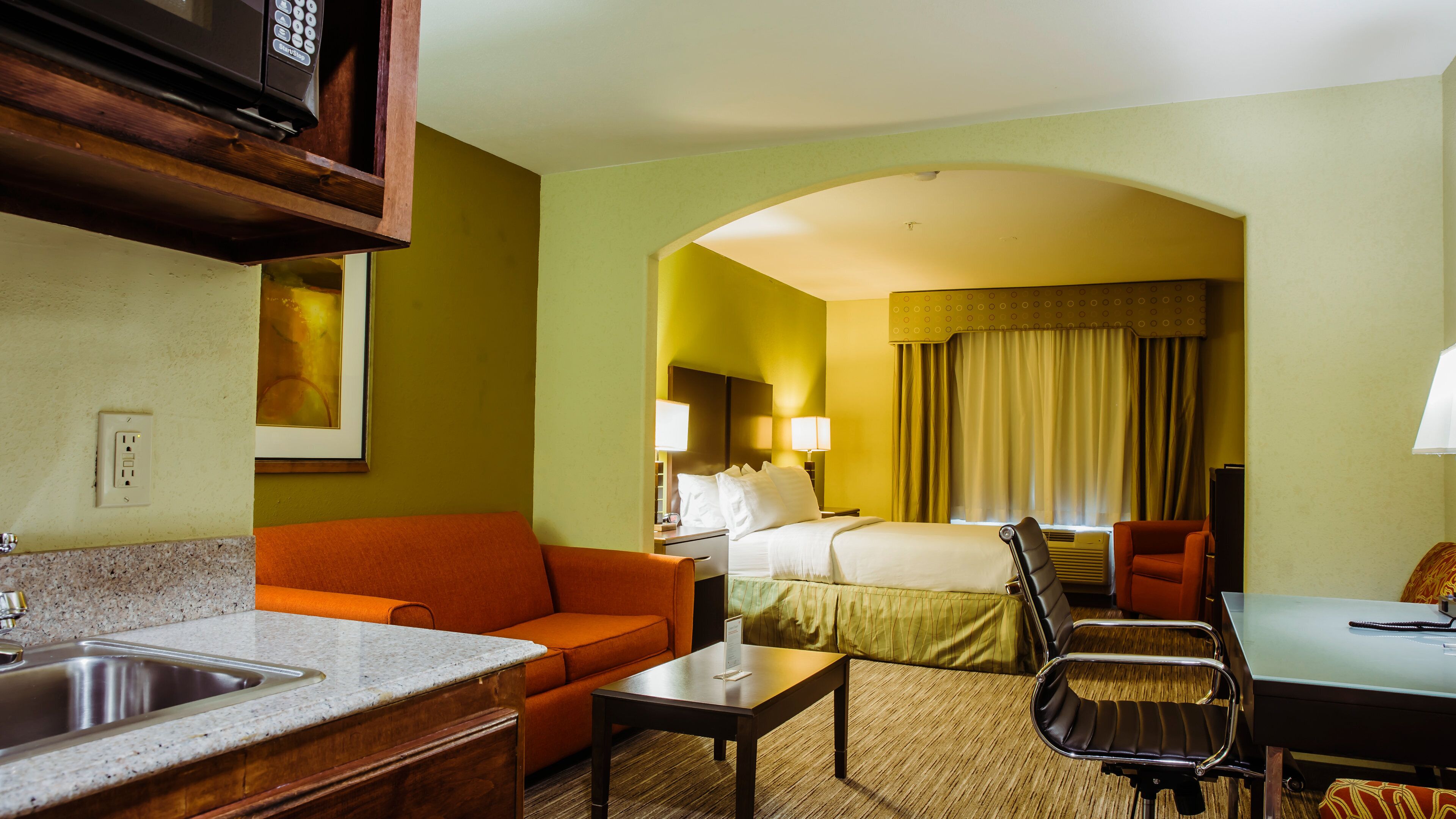 A hotel room with a kitchenette, a bed with a headboard, a sitting area with a sofa and armchair, and a dining table.