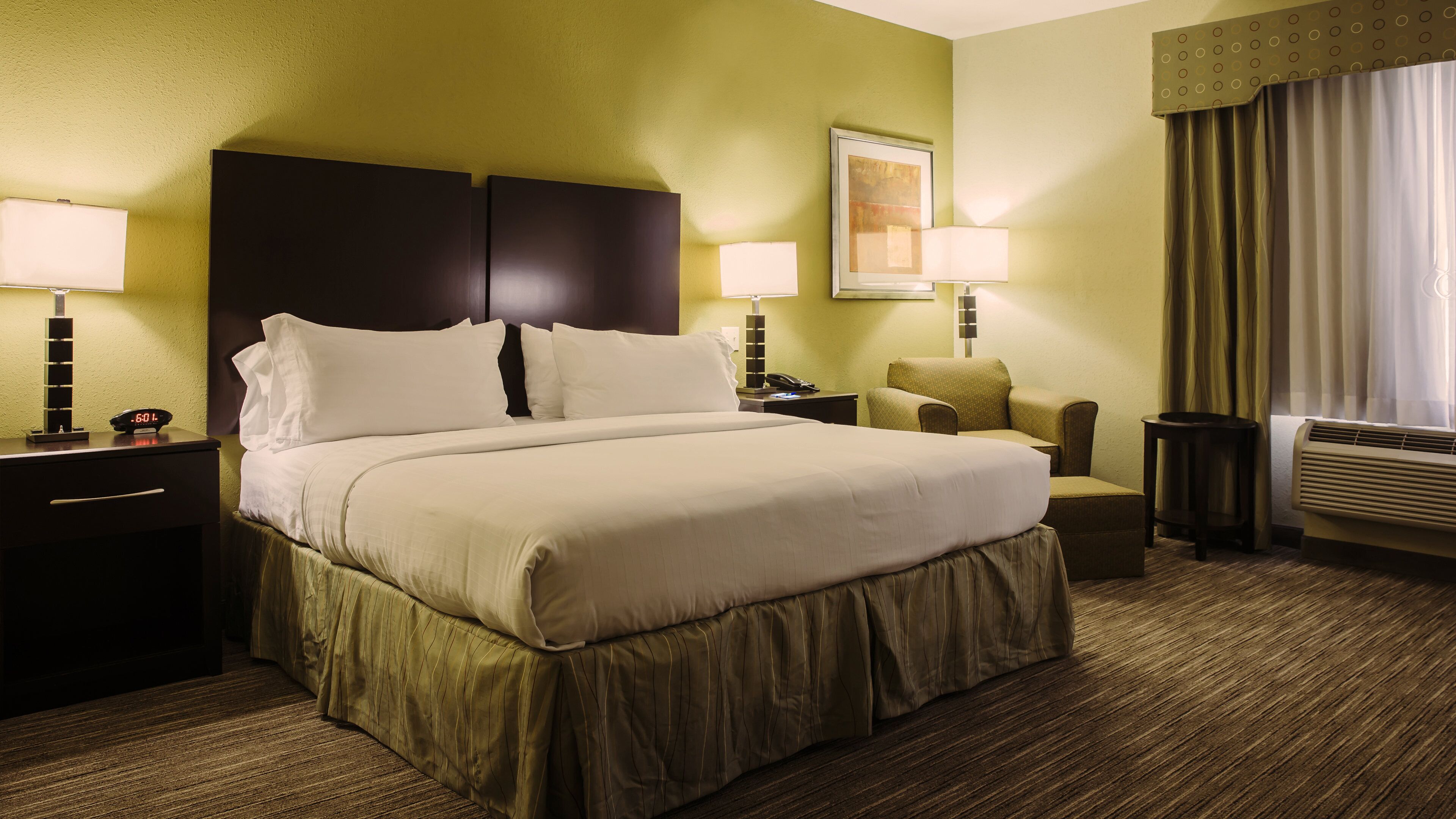 A hotel room with a large bed, two bedside tables, a chair, a desk, a lamp, a clock, and a window with curtains.