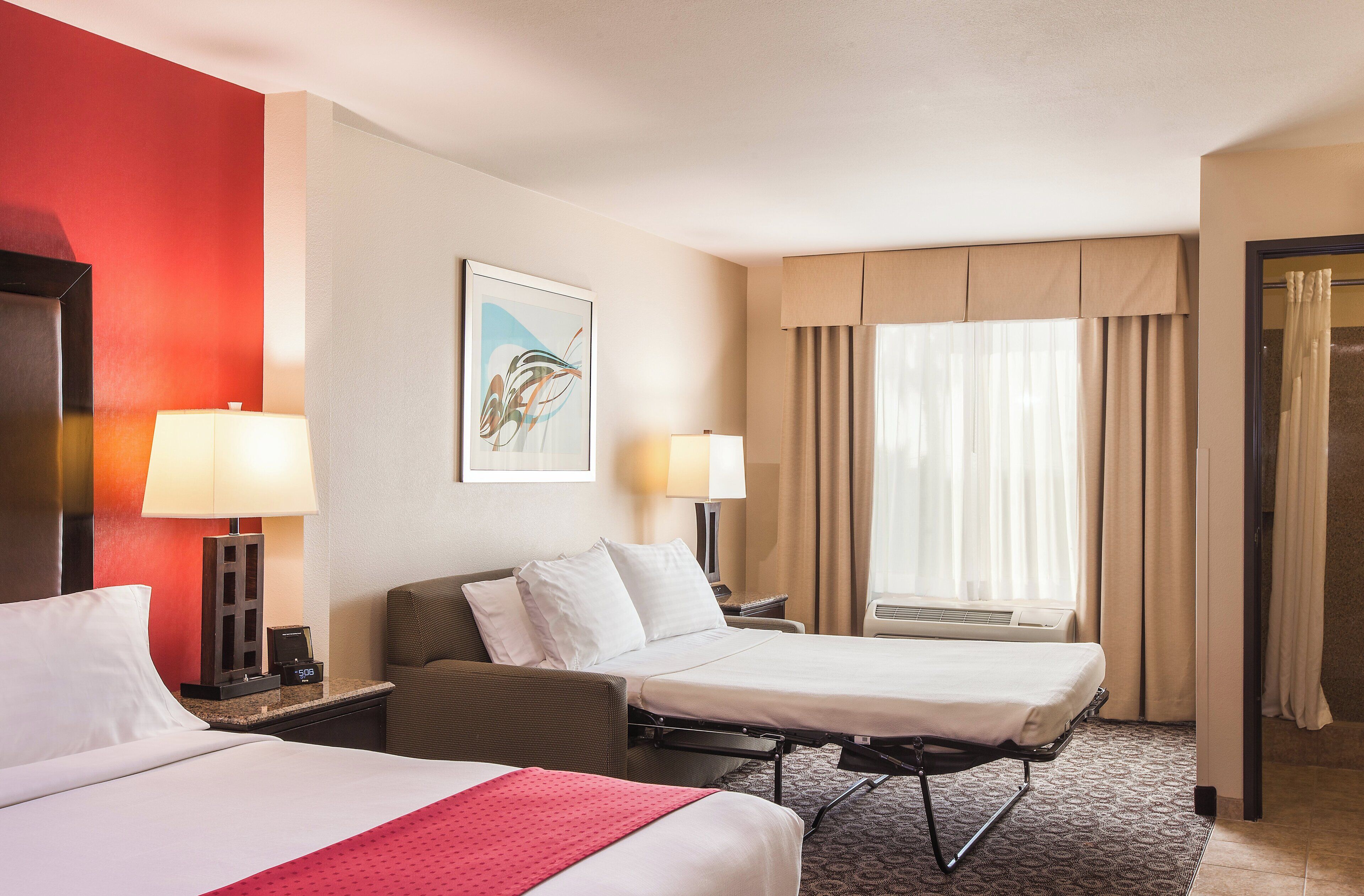 A hotel room with two beds, a desk, a chair, a lamp, a painting, and a window with curtains.