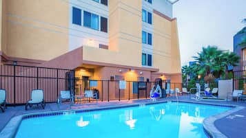 Área de piscina de hotel com piscina azul cristalina, espreguiçadeiras e um edifício com várias janelas e varandas.
