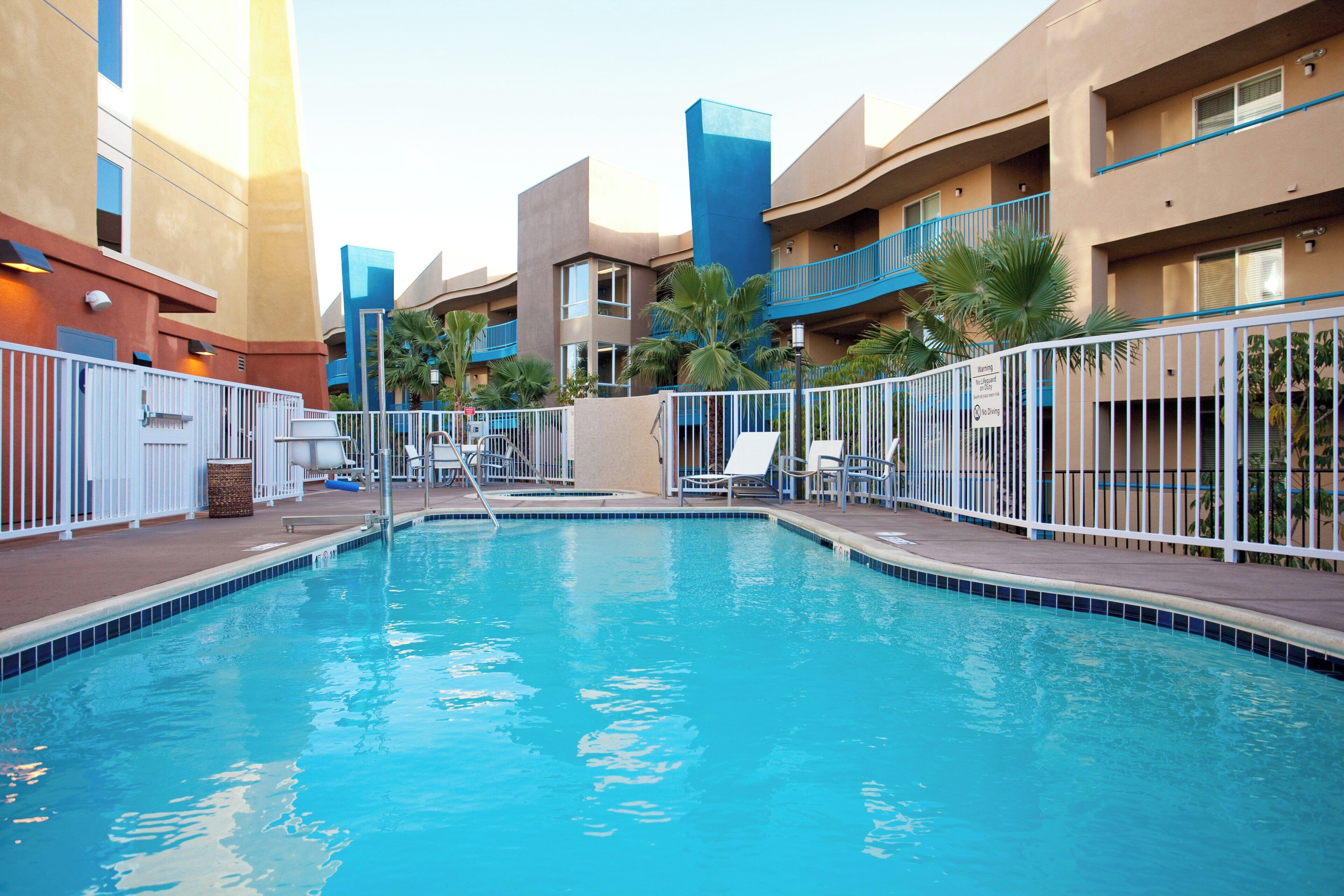 A swimming pool surrounded by a white fence, with lounge chairs and a palm tree.